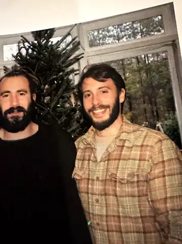 Jim Baker (left, in a black shirt) and his younger brother David Baker (right, in a plaid shirt) smiling side by side by a window