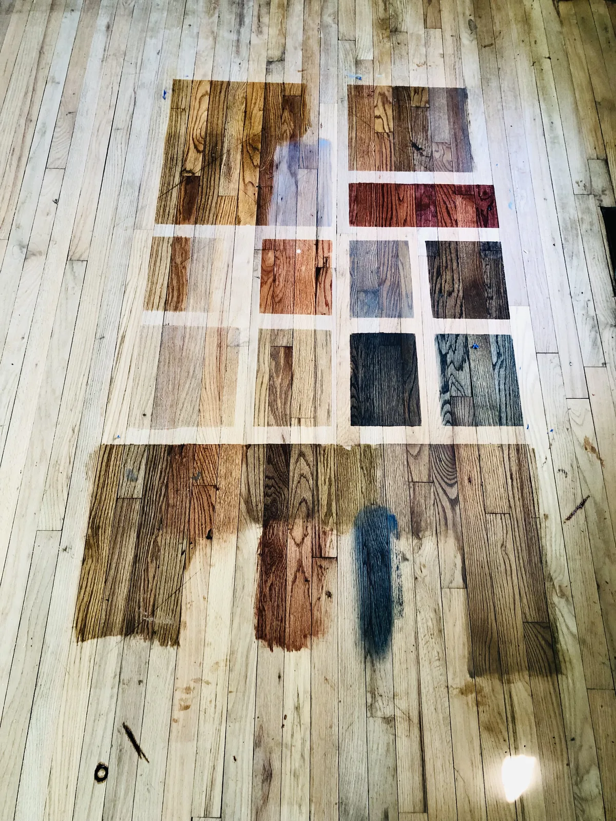 A grid of taped stain color samples laid directly on a bare oak floor