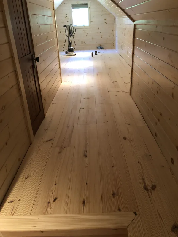 Attic room clad in light pine planking on sloped ceiling, walls and floor with a small window
