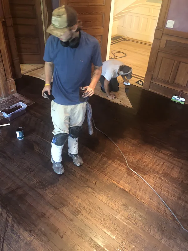 Two craftsmen kneeling to apply filler to a freshly stained brown plank floor near wood wainscot