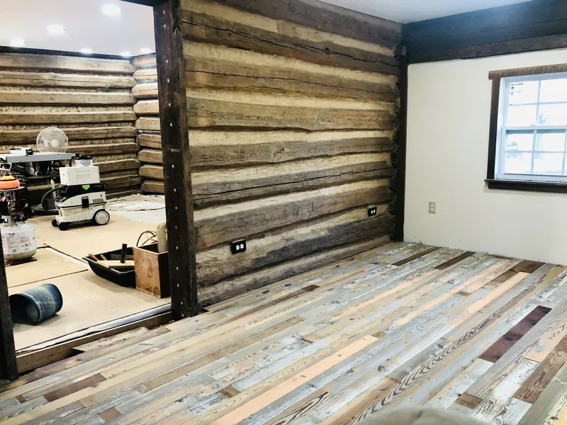 Room mid-installation with mixed reclaimed floorboards and an exposed chinked log cabin wall