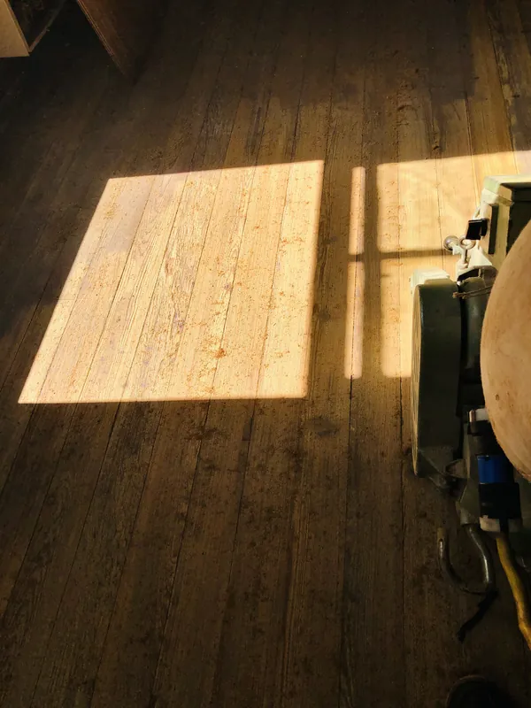 Dark aged plank floor with a bright sunlit patch and a floor buffer machine at right in warm low light
