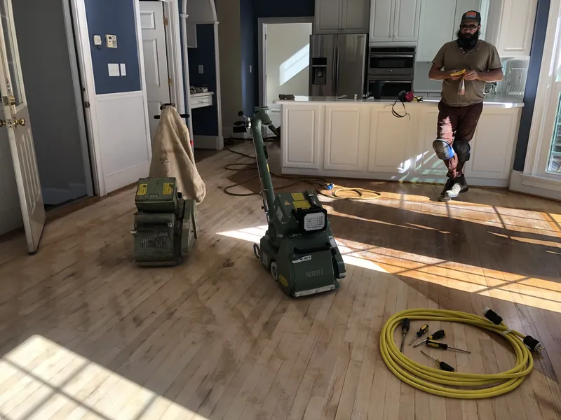 Two drum sanders and a power cord on bare oak floor with a craftsman in the kitchen beyond