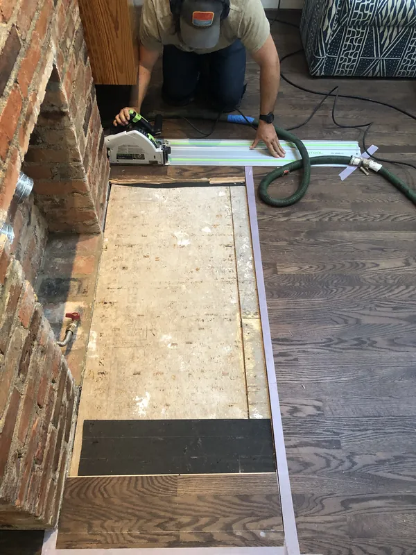 Craftsman nailing light oak flooring beside an exposed brick fireplace foundation
