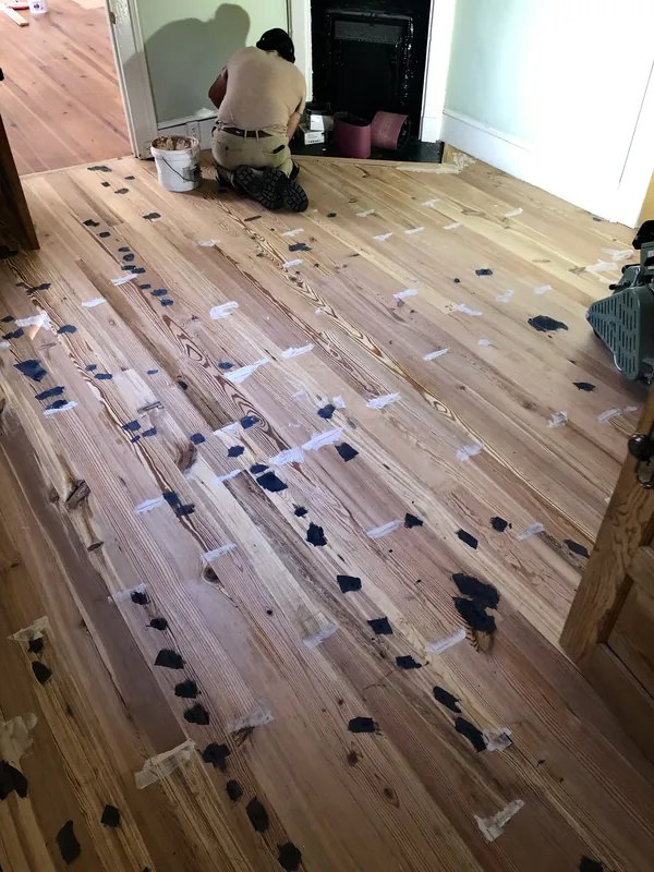 Craftsman kneeling to trowel dark filler into sanded heart pine planks dotted with putty patches