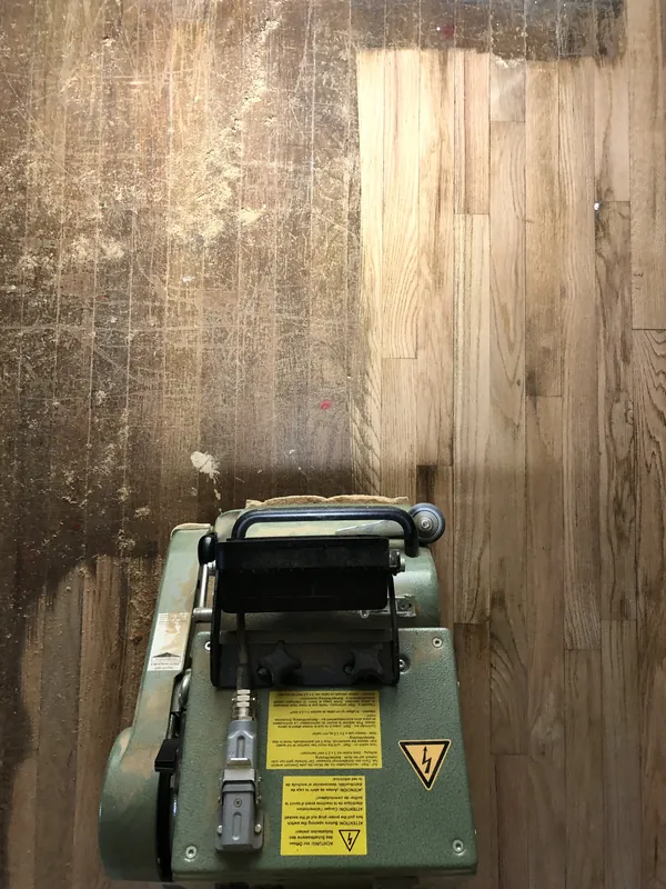 Drum sander on an oak floor showing a lighter sanded strip against worn darker finish