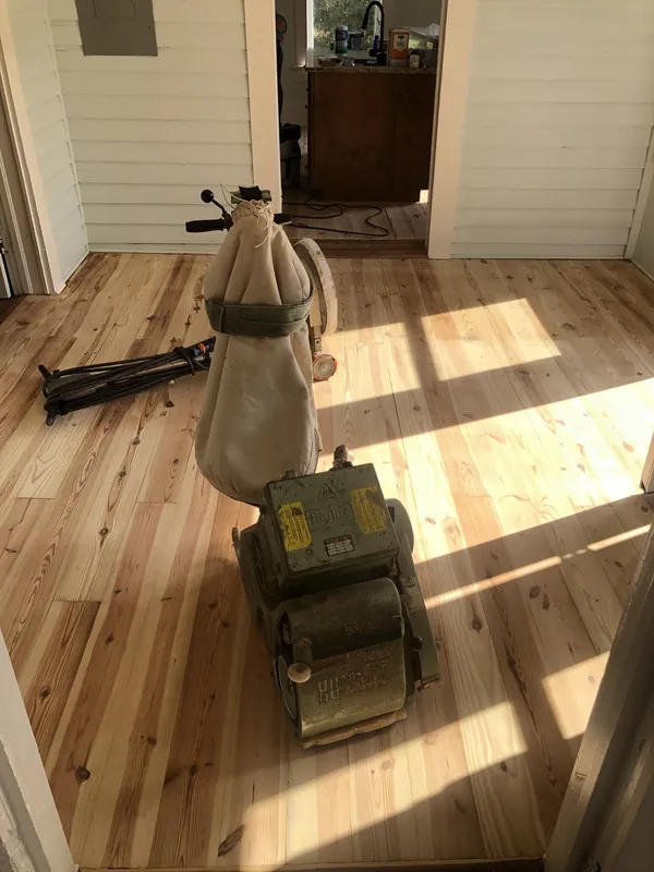 Belt drum sander with dust bag mid-pass across light pine flooring in raking sunlight