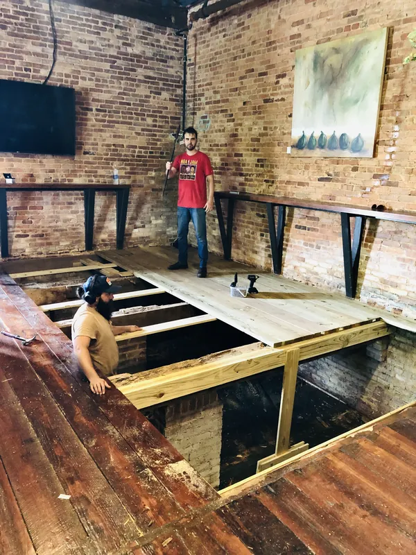 Two craftsmen installing floor joists and decking over an open cavity in a brick-walled room