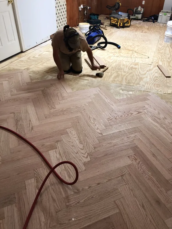 Craftsman kneeling to install red oak herringbone flooring with tools in the background