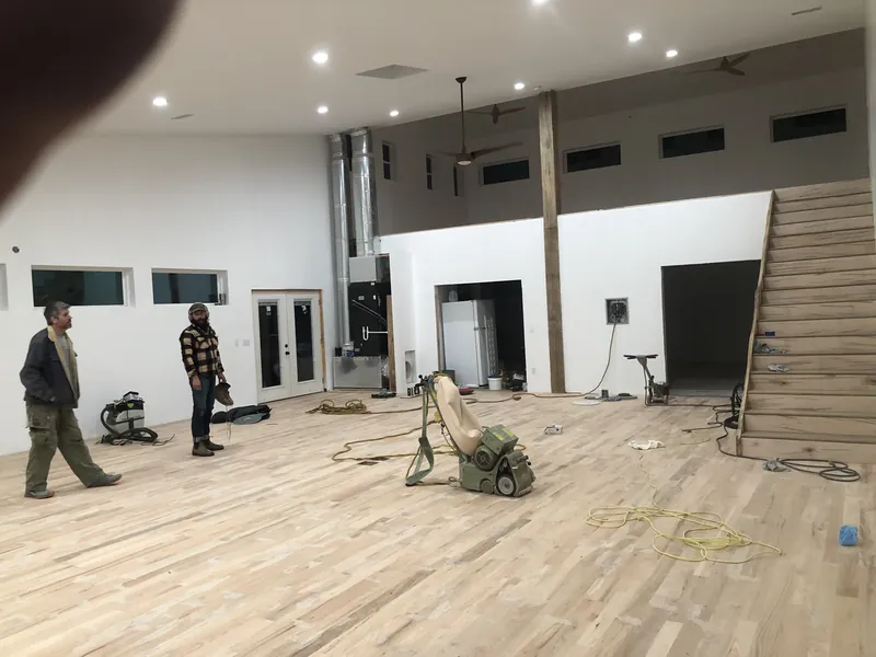 Large open room mid-install with sander, cordage, a staircase and two craftsmen