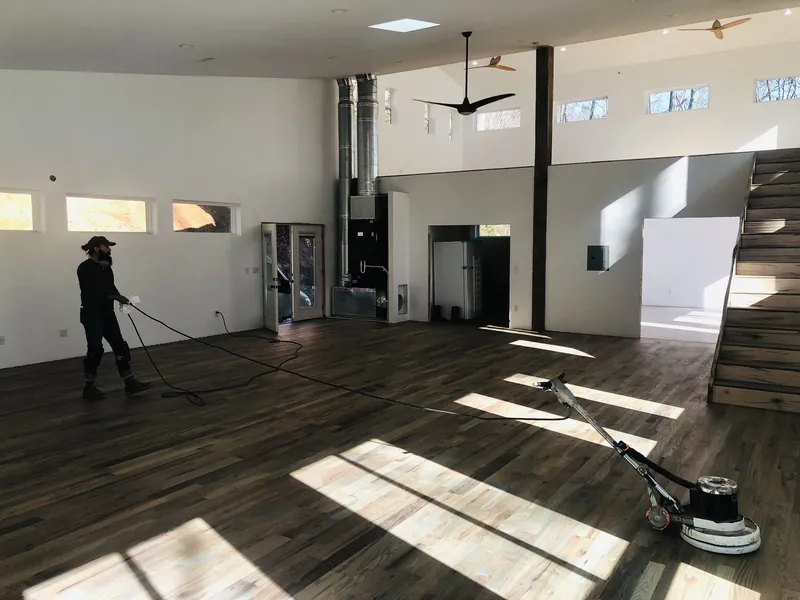 Craftsman running a floor buffer across pine flooring in a sunlit open room with tall walls and open stairs